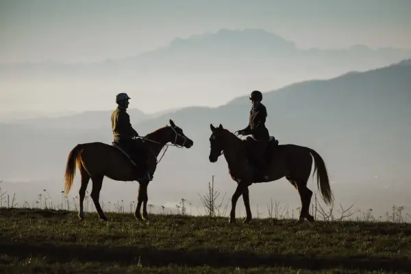 Due cavalieri in silhouette godono di una **passeggiata a cavallo in Lombardia** al tramonto, con montagne nebbiose all'orizzonte.