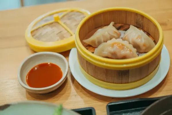 Parujące pierogi gyoza w bambusowym parowniku, obok miseczka z sosem.