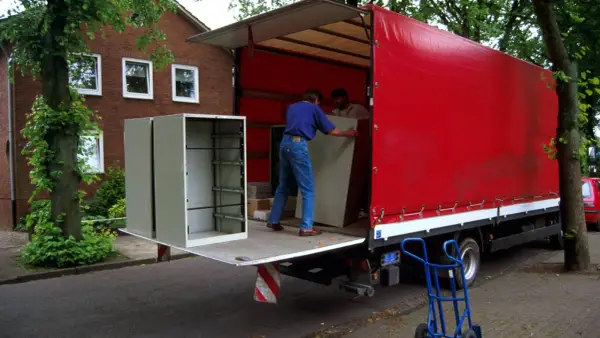 Umzugshelfer laden einen Schrank auf einen roten LKW. Der Schrank steht am Straßenrand, was die Bedeutung von "kerbside" verdeutlicht.