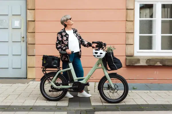 Ältere Dame mit QIO Bike, positive Erfahrungen mit dem E-Bike, das mit einem Helm und einer Tasche beladen ist.