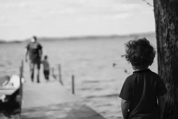 Bambino con capelli ricci guarda verso un molo sfocato dove camminano due figure. Un senso di isolamento, quasi un attaccamento disorganizzato, aleggia nell'aria.