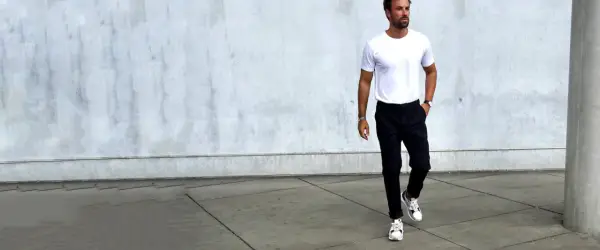 Mann in weißem T-Shirt und schwarzer Chino-Hose, dazu weiße Sneaker. Ein Beispiel, welche Schuhe zur Chino Hose Herren passen.