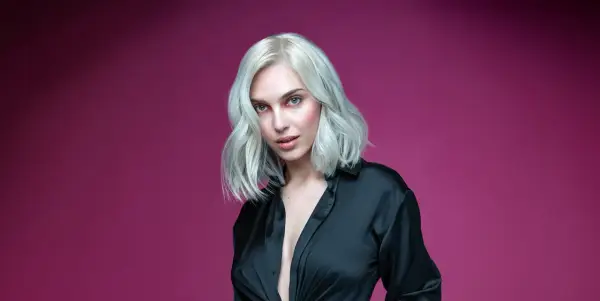 Mujer con cabello rubio ceniza platino, ojos azules y camisa negra abierta, sobre fondo rosa.