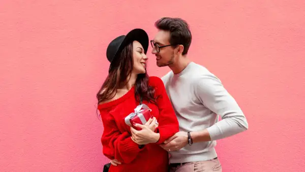 Para zakochanych na tle różowej ściany. Ona trzyma prezent, on obejmuje ją ramieniem. Mówi mu "I love you" po angielsku.