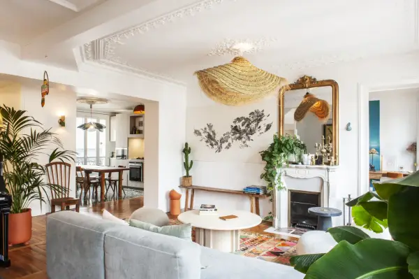 Salon au style classique revisité, avec un canapé gris, un tapis coloré, une cheminée ornée et des plantes luxuriantes.