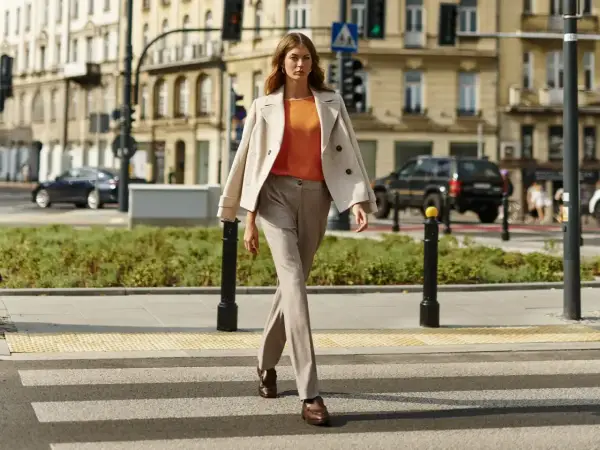 Elegancka kobieta w beżowym płaszczu i pomarańczowej bluzce przechodzi przez pasy. Stylowe stylizacje do beżowego płaszcza na co dzień.
