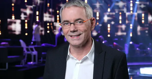 Uśmiechnięty Robert Janowski w okularach na tle sceny. Wiele osób zastanawia się, ile lat ma Robert Janowski, ale jego wiek nie jest widoczny na zdjęciu.