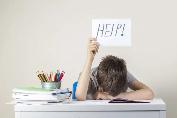 Chłopiec z kartką "HELP!" nad głową, przy biurku pełnym książek i kredek. Może to być sygnał trudności, wymagający diagnozy psychologicznej.