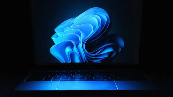 Niebieski, falujący kształt na ekranie laptopa z podświetlaną klawiaturą, przypominający logo Windows 11 24H2.
