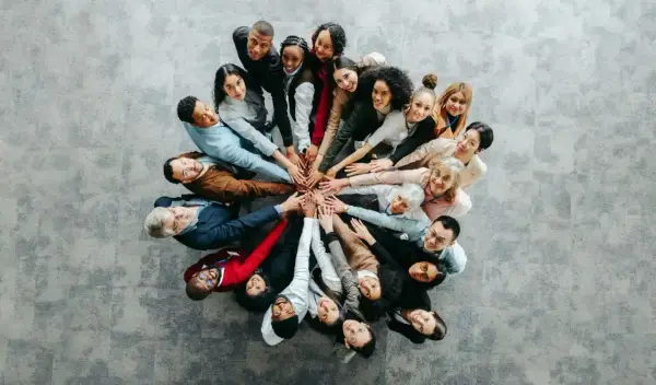 Grupa ludzi w kręgu, dłonie złożone na środku, symbolizująca współpracę przy wypełnianiu kwestionariusza osobowego dla osoby ubiegającej się o zatrudnienie.