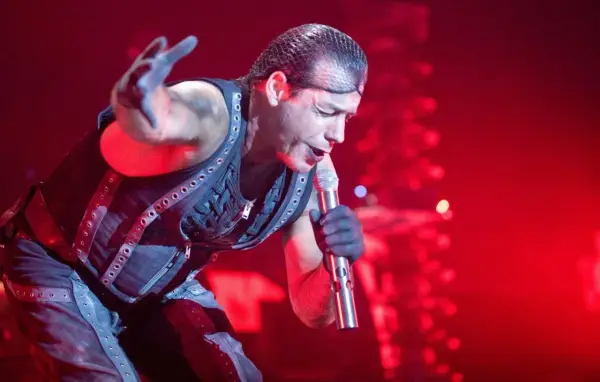 Till Lindemann, chanteur du groupe metal allemand Rammstein, en pleine performance sur scène, micro à la main, sous un éclairage rouge intense.