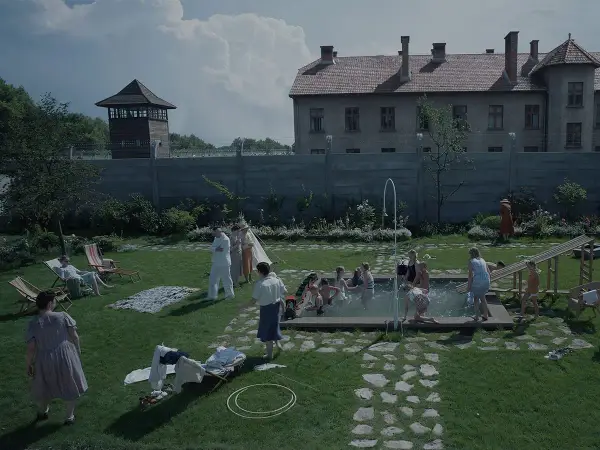 Scena da film strani: bambini giocano in piscina, adulti sorvegliano, mentre sullo sfondo incombono torri di guardia e un edificio imponente.