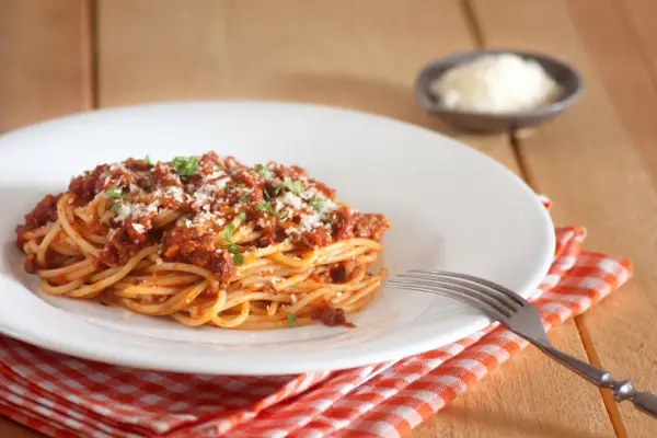 Talerz spaghetti bolognese z parmezanem i natką pietruszki, obok widelec i ser w miseczce.