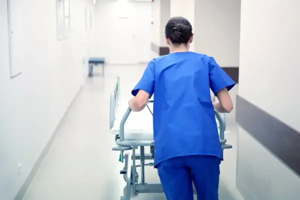 Salowa w niebieskim uniformie pcha łóżko szpitalne korytarzem. To część jej obowiązków w szpitalu.