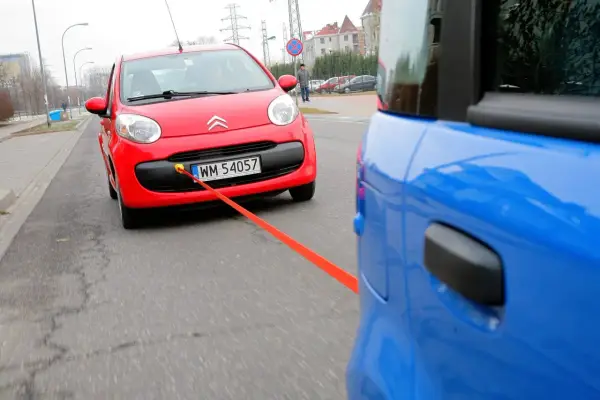 Czerwony Citroen holowany przez niebieski samochód. Pomarańczowa linka holownicza jest napięta, sugerując ruch i prędkość.