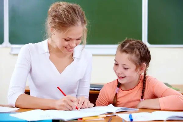 Uśmiechnięta nauczycielka wspomagająca z kwalifikacjami pomaga uczennicy w lekcjach przy biurku.