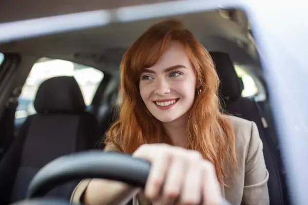 Donna sorridente al volante, con capelli rossi e lentiggini. Pensa alla sua assicurazione giornaliera auto per un viaggio sereno.