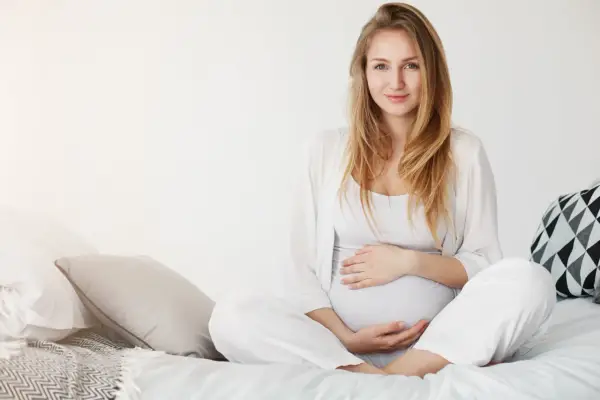 Uśmiechnięta kobieta w ciąży w białych ubraniach siedzi na łóżku, delikatnie trzymając się za brzuch.