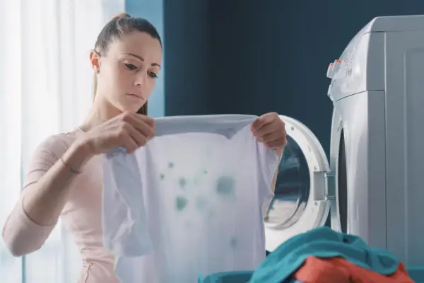 Zmartwiona kobieta trzyma białą koszulkę z zielonymi plamami, która wygląda jak zafarbowane ubrania. Obok stoi otwarta pralka.