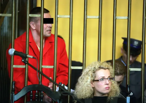 Mężczyzna w czerwonym ubraniu, przypominający lelek gangster, za kratami. Obok kobieta w okularach i policjant.
