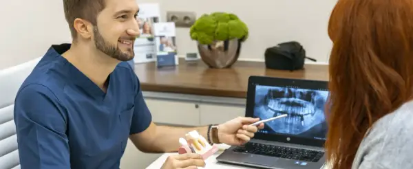 Dentysta omawia z pacjentką plan leczenia, pokazując na ekranie zdjęcie rentgenowskie szczęki i model zębów, wyjaśniając znaczenie czyszczenia dziąseł.