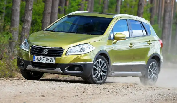 Zielony Suzuki S-Cross na drodze leśnej, wzbudzający pozytywne suzuki s-cross opinie. Kurz spod kół świadczy o jego terenowych możliwościach.