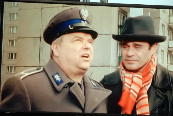 Dwóch mężczyzn w kapeluszach, jeden w mundurze, drugi w szaliku. Brunet wieczorową porą, w czarnym kapeluszu i czerwonym szaliku, rozmawia z policjantem.