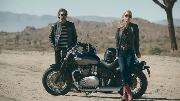 Para w skórzanych kurtkach pozuje przy motocyklu Triumph Speedmaster na pustynnym tle.