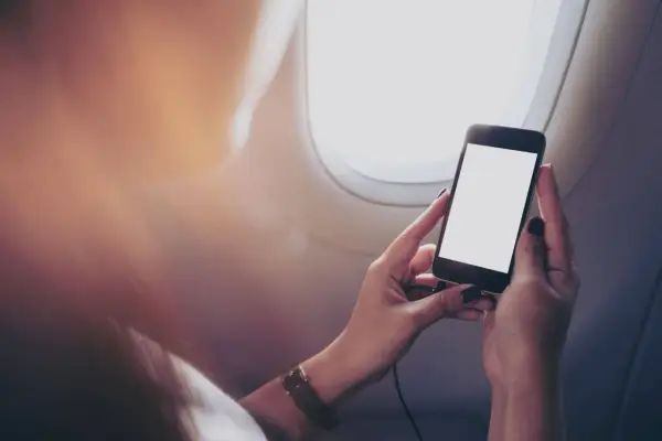 Kobieta trzyma telefon przy oknie w samolocie. Czy w samolocie można korzystać z telefonu? Tak, ale z ograniczeniami.
