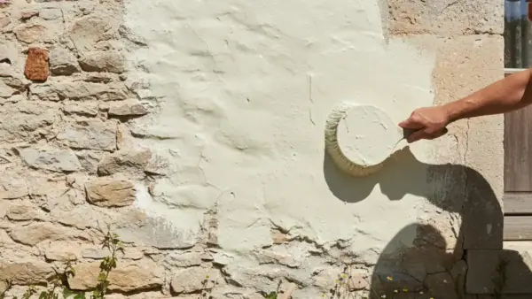 Une main applique une peinture claire sur un mur en pierre enduit de chaux. La question "peut on peindre sur de la chaux" trouve ici sa réponse visuelle.