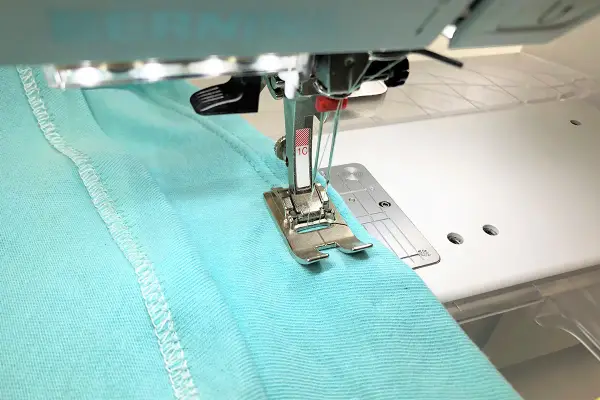A double needle stitches a hem on turquoise fabric. Most sewing machines can use a double needle with the right setup.
