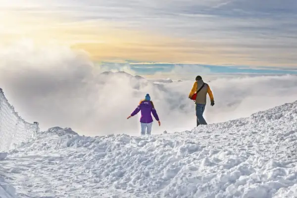 Dwie osoby wędrują po ośnieżonym szczycie. W oddali widać góry i chmury. Czyżby szukali odpowiedzi na pytanie: jakie raki w Tatry?