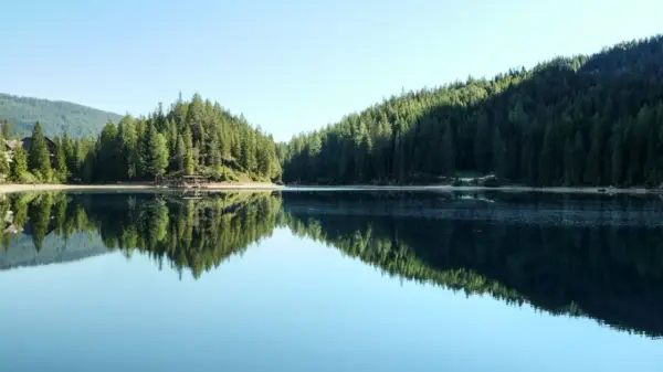 Spokojna tafla jeziora odbija gęsty las i góry. Widok na malowniczą kotlinę z drewnianym pomostem.