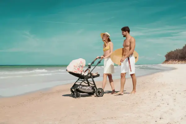 Para spaceruje po plaży z wózkiem Kinderkraft Askoy. Mężczyzna niesie deskę surfingową.