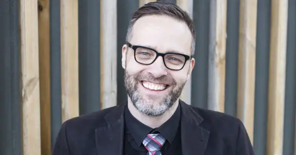 A smiling man with glasses and a beard, wearing a suit and tie, stands in front of a wooden slatted background.