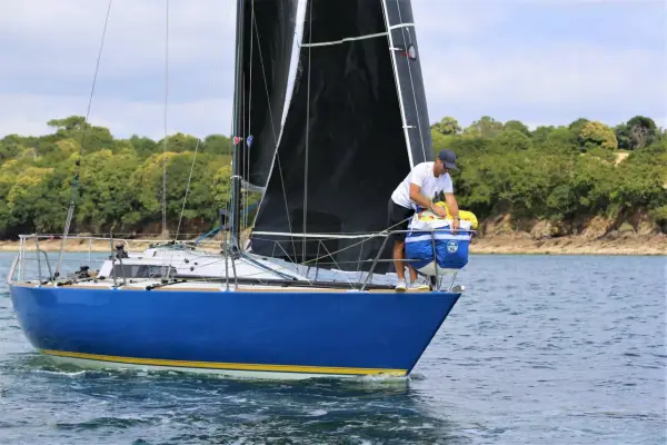 Un homme prépare le bateau, une voile noire déployée, sur fond de côte boisée. Il semble être le **half toner** de l'équipage.