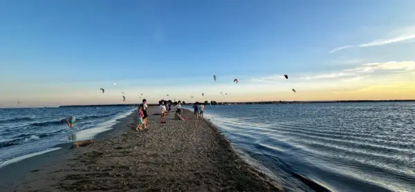 Ludzie spacerują po piaszczystej mierzei, podczas gdy na niebie widać liczne latawce do kitesurfingu.