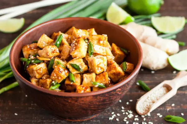 Pokrojone w kostkę, glazurowane tofu z zieloną cebulką i sezamem w misce. Obok limonka, imbir i drewniana łyżka z sezamem.