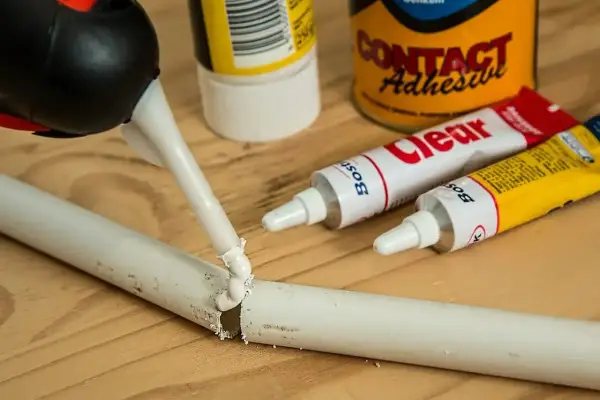 Pistolet z klejem aplikuje masę na pękniętą białą rurkę. Obok leżą tubki kleju, pokazujące, czym skleić plastik.
