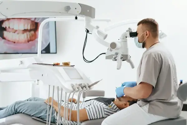 Stomatolog w masce i rękawiczkach bada pacjenta pod mikroskopem. Na ekranie widać zbliżenie zębów.