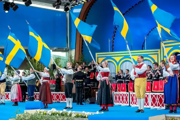 Människor i folkdräkter viftar svenska flaggor under firandet av 6 juni i Stockholm. En orkester spelar i bakgrunden.