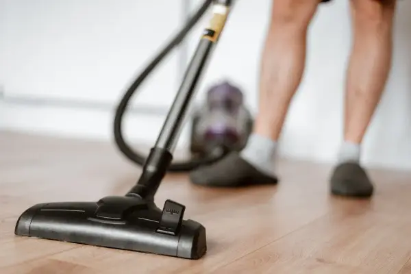 Odkurzacz na drewnianej podłodze, część nóg w kapciach i skarpetkach. To jeden z obowiązków domowych po angielsku – "vacuuming".