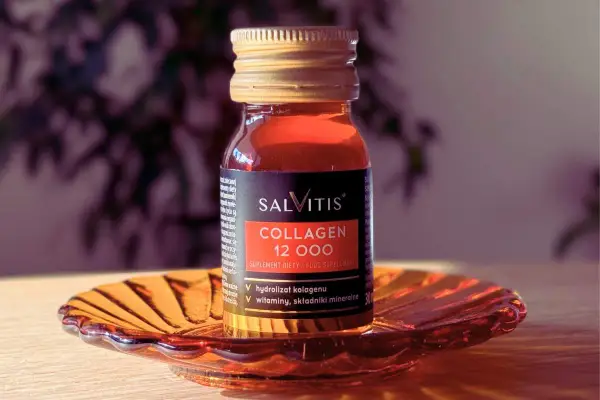 Szklana buteleczka suplementu diety SALVITIS Collagen 12 000, z płynem o bursztynowym kolorze, stoi na ozdobnej, pomarańczowej podstawce.