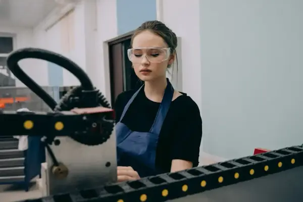 Młody pracownik produkcji w okularach ochronnych i fartuchu pracuje przy maszynie.