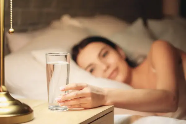 Une main attrape un verre d'eau posé à droite sur une table de chevet, une femme se réveille en arrière-plan.