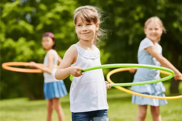 Uśmiechnięte dziewczynki biorą udział w konkurencjach sportowych dla dzieci, kręcąc hula-hop na trawie.
