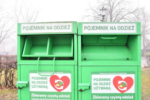 Dwa zielone kontenery na odzież używaną, z napisem "POJEMNIK NA ODZIEŻ UŻYWANĄ" i sercem z symbolami recyklingu.