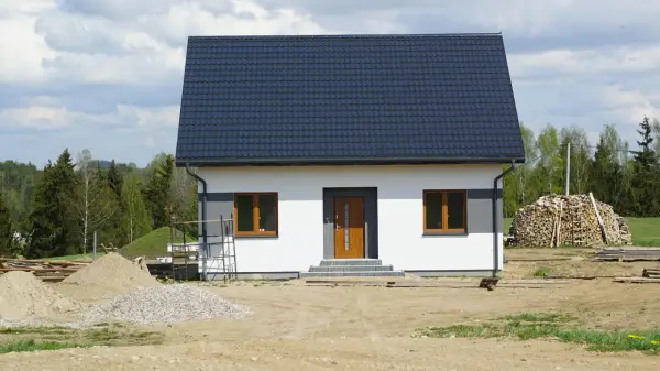 Nowy dom z ciemnym dachem i białymi ścianami, gotowy na zawiadomienie o zakończeniu budowy. Wokół plac budowy z piaskiem i drewnem.