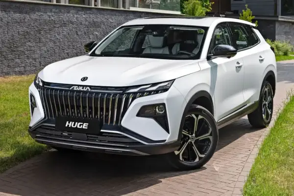 Biały, chiński SUV z imponującym grillem i napisem "HUGE" na masce, zaparkowany na podjeździe z kostki brukowej.