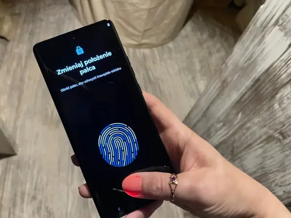 Dłoń z telefonem, na ekranie skaner linii papilarnych i komunikat "Zmieniaj położenie palca". To jak próba ucieczki z cyfrowego więzienia, niczym **jailbreak**.